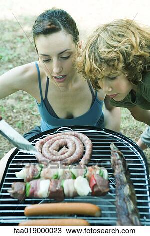 Stock Image - Young woman and boy bending over barbecue, looking at grilling meats. Fotosearch