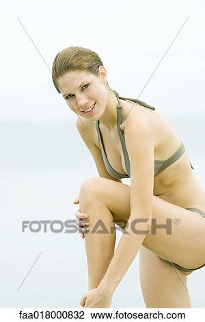 Young woman in bikini standing with one knee up, touching leg, smiling at camera View Large Photo Image Stock Image - Young woman in bikini standing with one knee up, touching leg, smiling at camera. Fotosearch