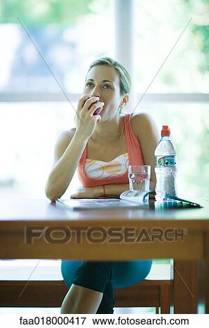 Stock Photo - Young woman in exercise clothes sitting with bottle of water and magazine, eating apple. Fotosearch