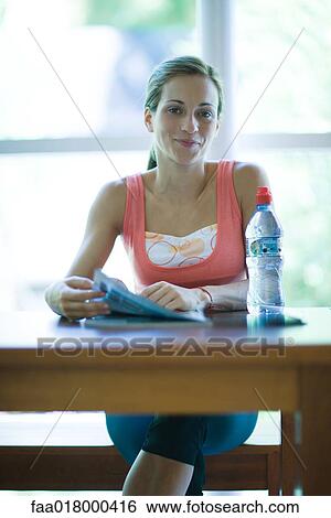 Stock Photograph - Young woman in exercise clothes sitting with bottle of water and magazine, smiling at camera. Fotosearch