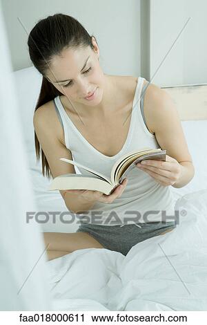 Stock Image - Young woman sitting on bed, reading book. Fotosearch