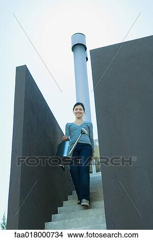 Picture - Young woman walking down stairs, carrying watering can. Fotosearch