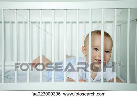 Baby lying on stomach, looking through bars of crib, smiling View Large Photo Image Stock Photograph - Baby lying on stomach, looking through bars of crib, smiling. Fotosearch