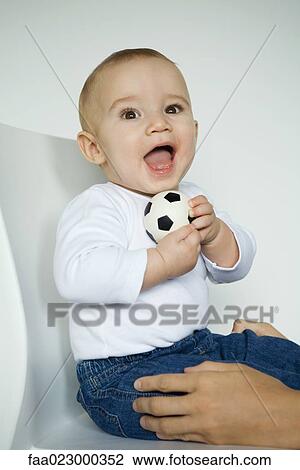 Stock Image - Baby sitting in chair holding ball, mother's hands holding baby's legs. Fotosearch