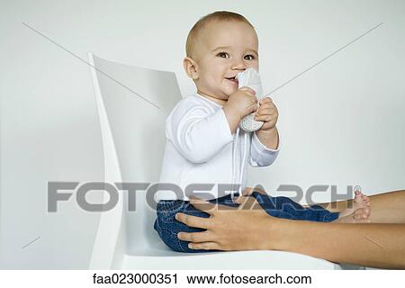 Baby sitting in chair putting shoe in mouth, mother's arms holding baby's legs View Large Photo Image Stock Image - Baby sitting in chair putting shoe in mouth, mother's arms holding baby's legs. Fotosearch