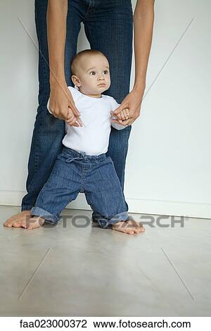 Stock Image - Baby standing between mother's legs, looking away. Fotosearch