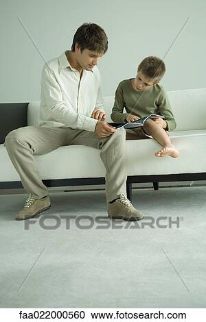 Stock Image - Boy and father sitting on sofa, looking at magazine. Fotosearch