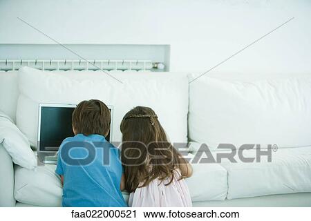 Stock Image - Boy and girl using laptop on sofa, rear view. Fotosearch