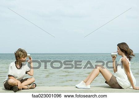 Stock Photograph - Boy and teenage girl sitting near ocean, talking through tin can phone. Fotosearch