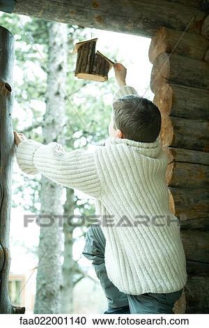 Stock Image - Boy in cabin window, reaching for birdhouse, rear view. Fotosearch