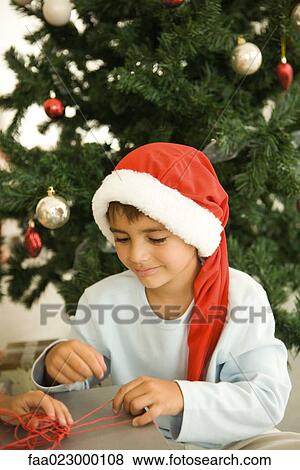 Stock Photo - Boy opening present in front of Christmas tree, wearing Santa hat, smiling. Fotosearch