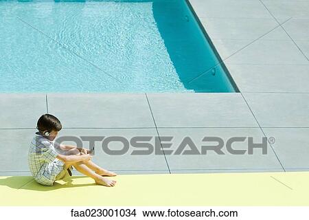 Boy sitting on the ground next to swimming pool, using handheld electronic device, wearing headphones View Large Photo Image Picture - Boy sitting on the ground next to swimming pool, using handheld electronic device, wearing headphones. Fotosearch