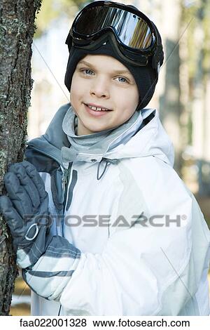Stock Photo - Boy wearing ski gear, leaning against tree trunk, smiling at camera, portrait. Fotosearch