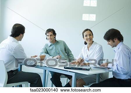 Stock Image - Business associates seated at table, chatting, one smiling at camera. Fotosearch
