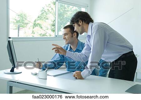 Businessman sitting at desk facing monitor while colleague stands next to him, speaking and pointing to screen View Large Photo Image Stock Photograph - Businessman sitting at desk facing monitor while colleague stands next to him, speaking and pointing to screen. Fotosearch