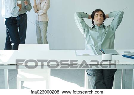 Businessman sitting at desk with hands behind head and eyes closed, colleagues standing in background View Large Photo Image Stock Photo - Businessman sitting at desk with hands behind head and eyes closed, colleagues standing in background. Fotosearch
