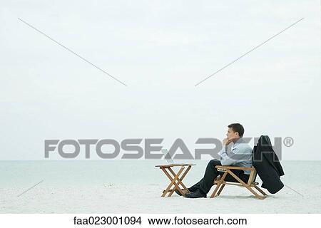 Businessman sitting on beach, looking at laptop computer, hand under chin, full length View Large Photo Image Picture - Businessman sitting on beach, looking at laptop computer, hand under chin, full length. Fotosearch