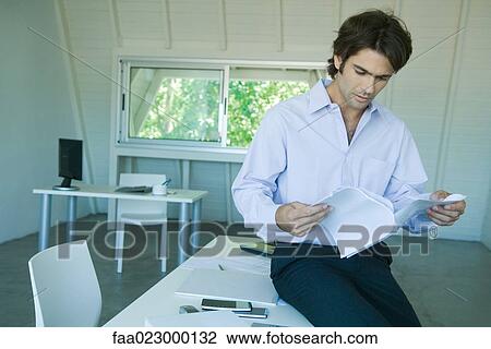 Businessman sitting on desk, reading document in office View Large Photo Image Stock Image - Businessman sitting on desk, reading document in office. Fotosearch