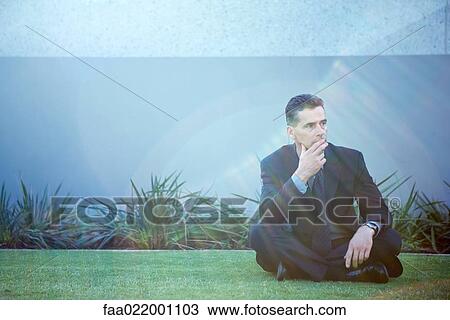 Stock Image - Businessman sitting on the ground outdoors, looking away, finger on lips, lens flare. Fotosearch