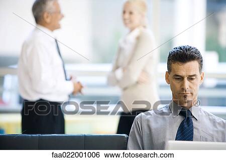 Stock Photograph - Businessman sitting, using laptop computer, associates standing in background. Fotosearch