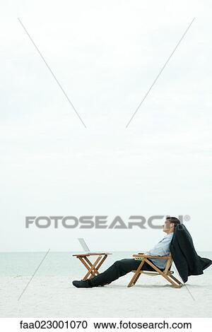 Stock Image - Businessman sitting with laptop computer at the beach, eyes closed. Fotosearch