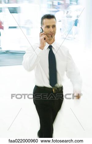 Stock Image - Businessman walking in lobby, using cell phone, blurred motion. Fotosearch
