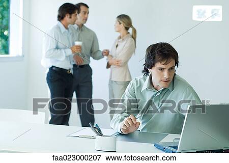 Businessman working on laptop, colleagues standing in background View Large Photo Image Stock Photo - Businessman working on laptop, colleagues standing in background. Fotosearch