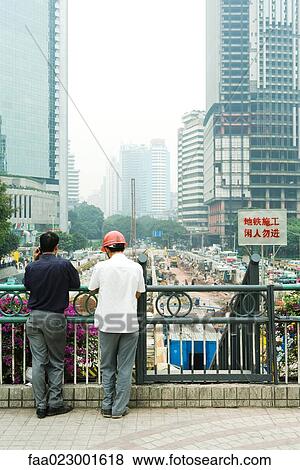 Stock Photo - China, Guangdong Province, Guangzhou, two men looking over construction site, rear view. Fotosearch
