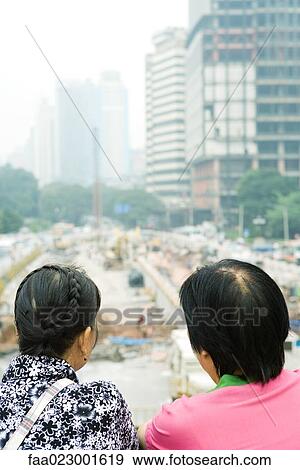 Stock Photo - China, Guangdong Province, Guangzhou, two people looking out over construction site, rear view, close-up. Fotosearch