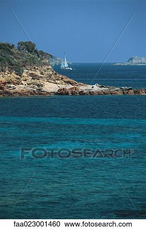 Stock Image - Corsica, sailboat sailing along coast, turquoise sea in foreground. Fotosearch