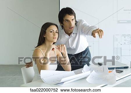 Stock Image - Couple next to table with blueprints, man leaning over woman's shoulder, pointing out of frame. Fotosearch