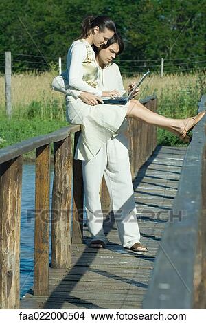 Picture - Couple on footbridge over water, woman sitting on rail, using laptop. Fotosearch