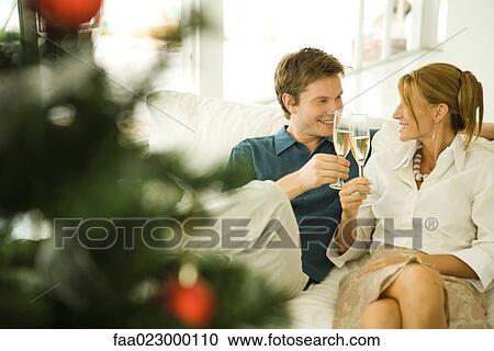 Stock Image - Couple sitting on couch, making a toast with champagne, smiling at each other. Fotosearch
