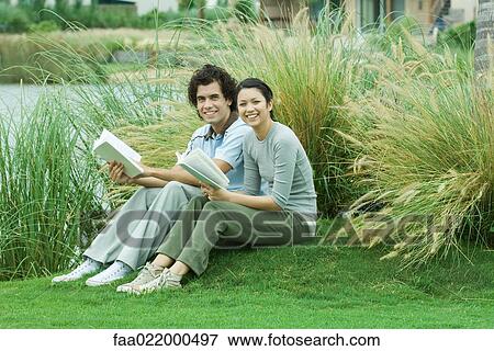 Couple sitting on grass with books, smiling at camera View Large Photo Image Stock Photo - Couple sitting on grass with books, smiling at camera. Fotosearch