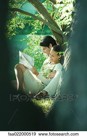 Couple sitting on ground, leaning against tree, reading newspaper, smiling, blurred tree trunk in foreground View Large Photo Image Stock Photo - Couple sitting on ground, leaning against tree, reading newspaper, smiling, blurred tree trunk in foreground. Fotosearch