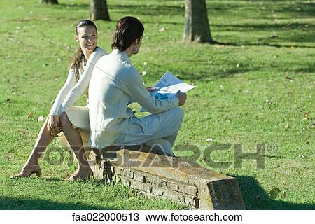 Stock Image - Couple sitting outdoors on low wall, man reading newspaper. Fotosearch