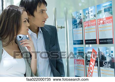 Stock Photography - Couple standing next to window of real estate agency, looking at for rent and for sale signs. Fotosearch