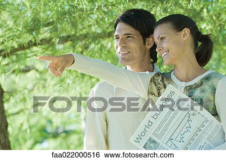 Couple standing outdoors holding newspaper, woman pointing, both looking out of frame View Large Photo Image Stock Photograph - Couple standing outdoors holding newspaper, woman pointing, both looking out of frame. Fotosearch