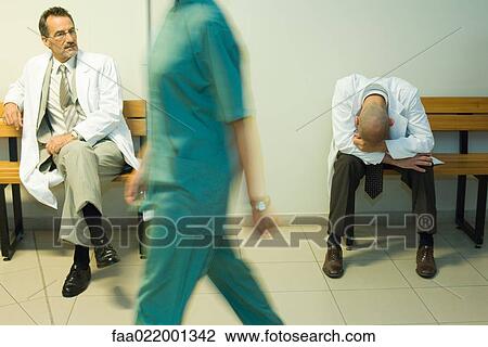 Stock Image - Doctors sitting in hospital corridor, one with head down. Fotosearch