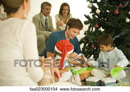 Father and two children sitting by Christmas tree, opening presents together, family watching View Large Photo Image Picture - Father and two children sitting by Christmas tree, opening presents together, family watching. Fotosearch