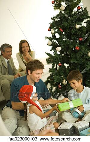 Father and two children sitting by Christmas tree, opening presents together, adults in background View Large Photo Image Stock Image - Father and two children sitting by Christmas tree, opening presents together, adults in background. Fotosearch