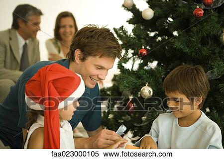 Stock Photography - Father and two children sitting by Christmas tree, opening presents together, daughter wearing Santa hat. Fotosearch
