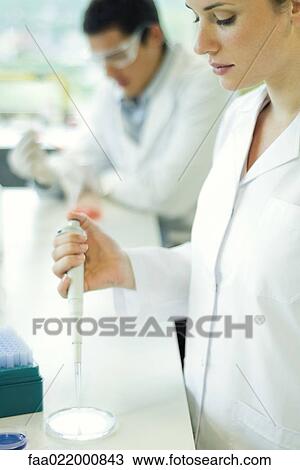 Stock Image - Female lab worker dropping solution into Petri dish. Fotosearch