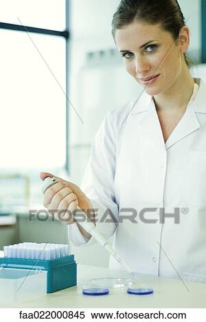 Stock Photography - Female lab worker dropping solution into Petri dish, smiling at camera. Fotosearch