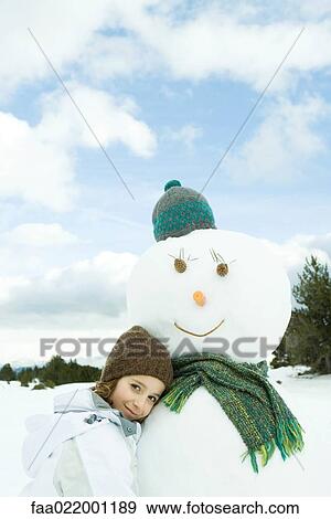 Stock Photo - Girl leaning against snowman, smiling at camera, portrait. Fotosearch