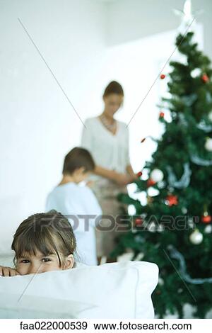 Girl looking over sofa at camera, boy and mother decorating Christmas tree in background View Large Photo Image Stock Photo - Girl looking over sofa at camera, boy and mother decorating Christmas tree in background. Fotosearch