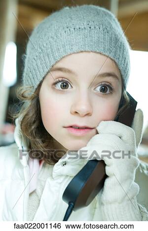 Stock Photograph - Girl using phone, dressed in winter clothing, looking away with wide eyes. Fotosearch
