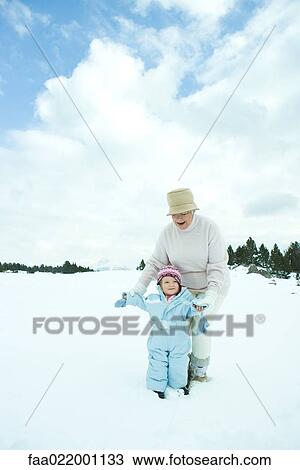 Stock Image - Grandmother and granddaughter standing together in snow, smiling, full length. Fotosearch