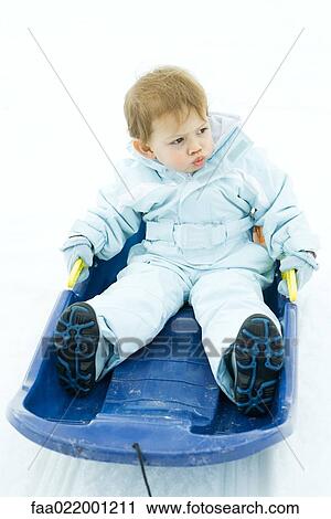 Stock Image - Little boy sitting on sled, looking away, frowning. Fotosearch