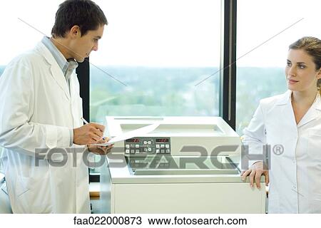 Stock Image - Male and female lab workers standing next to lab machine. Fotosearch